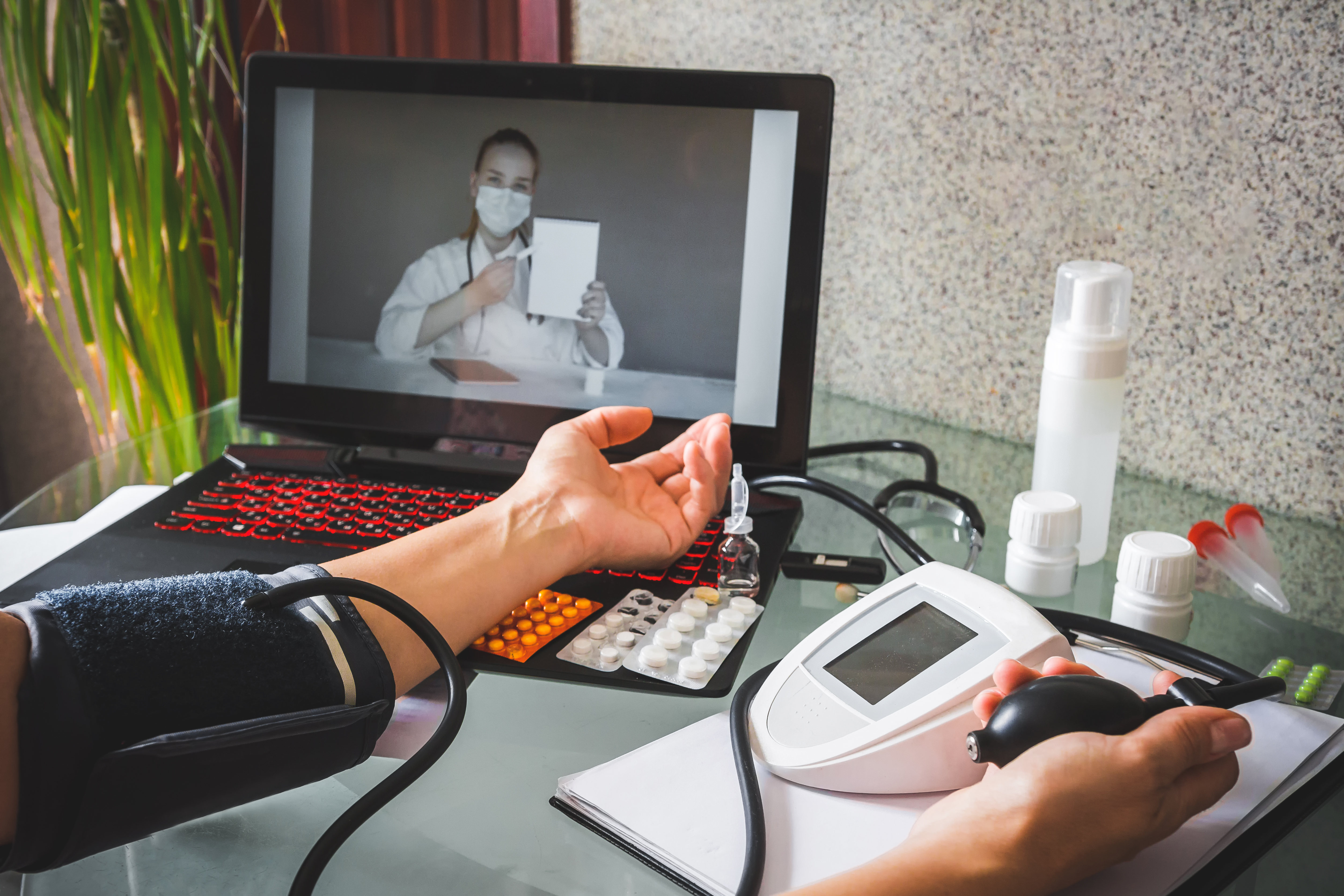 A doctor on a laptop screen observing a patient taking their blood pressure at home.