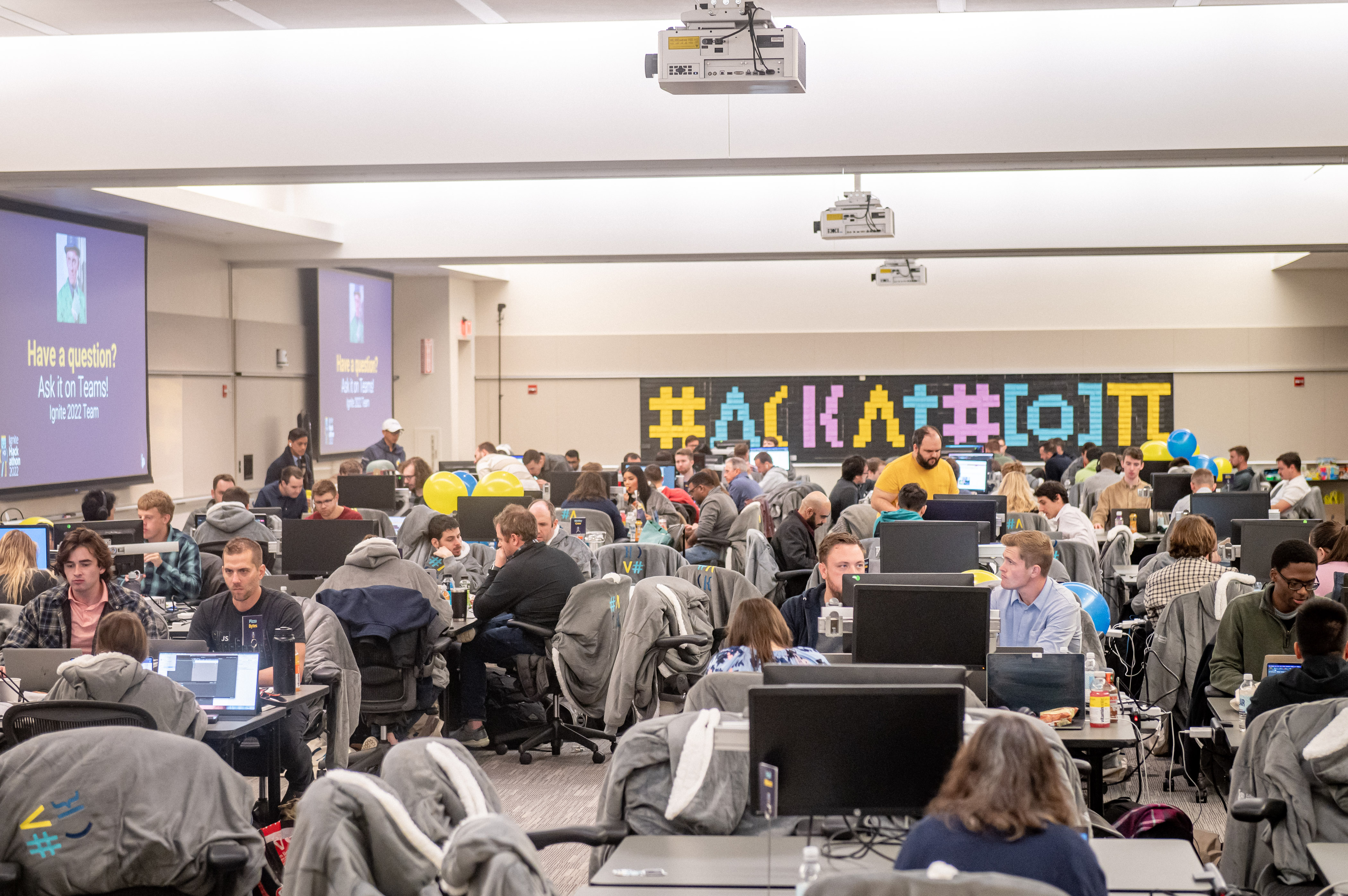 Large hall full of Liberty Mutual team members in groups with large monitors, the word “Hackathon” on a sign along one wall. 