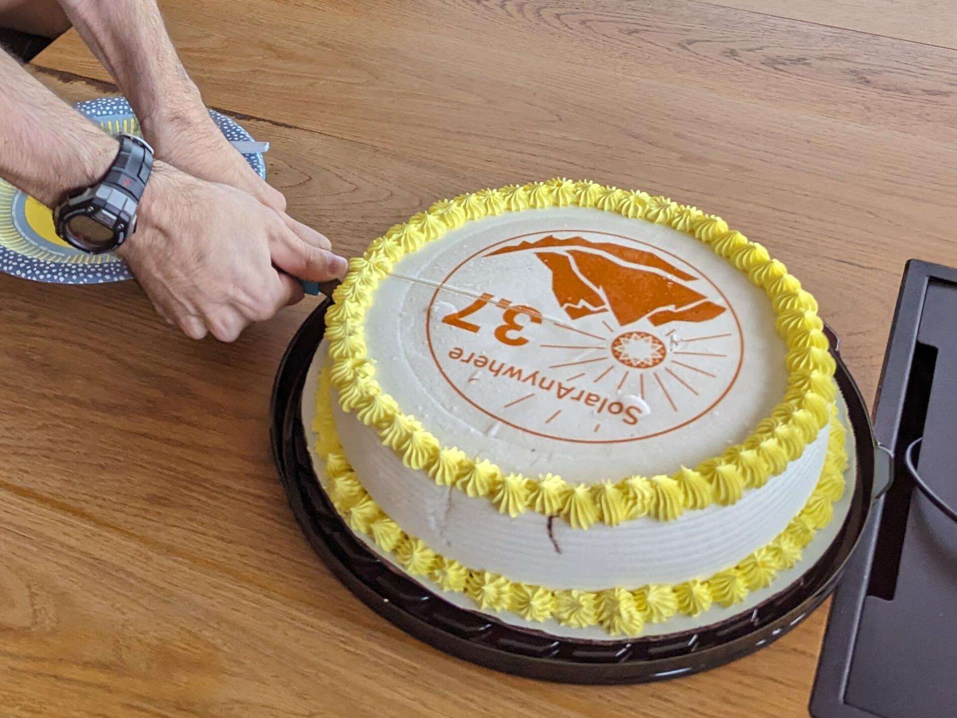 A cake reading SolarAnywhere 3.7 with a logo of mountains and a sunburst is cut by a person wearing a watch and holding the knife with both hands.