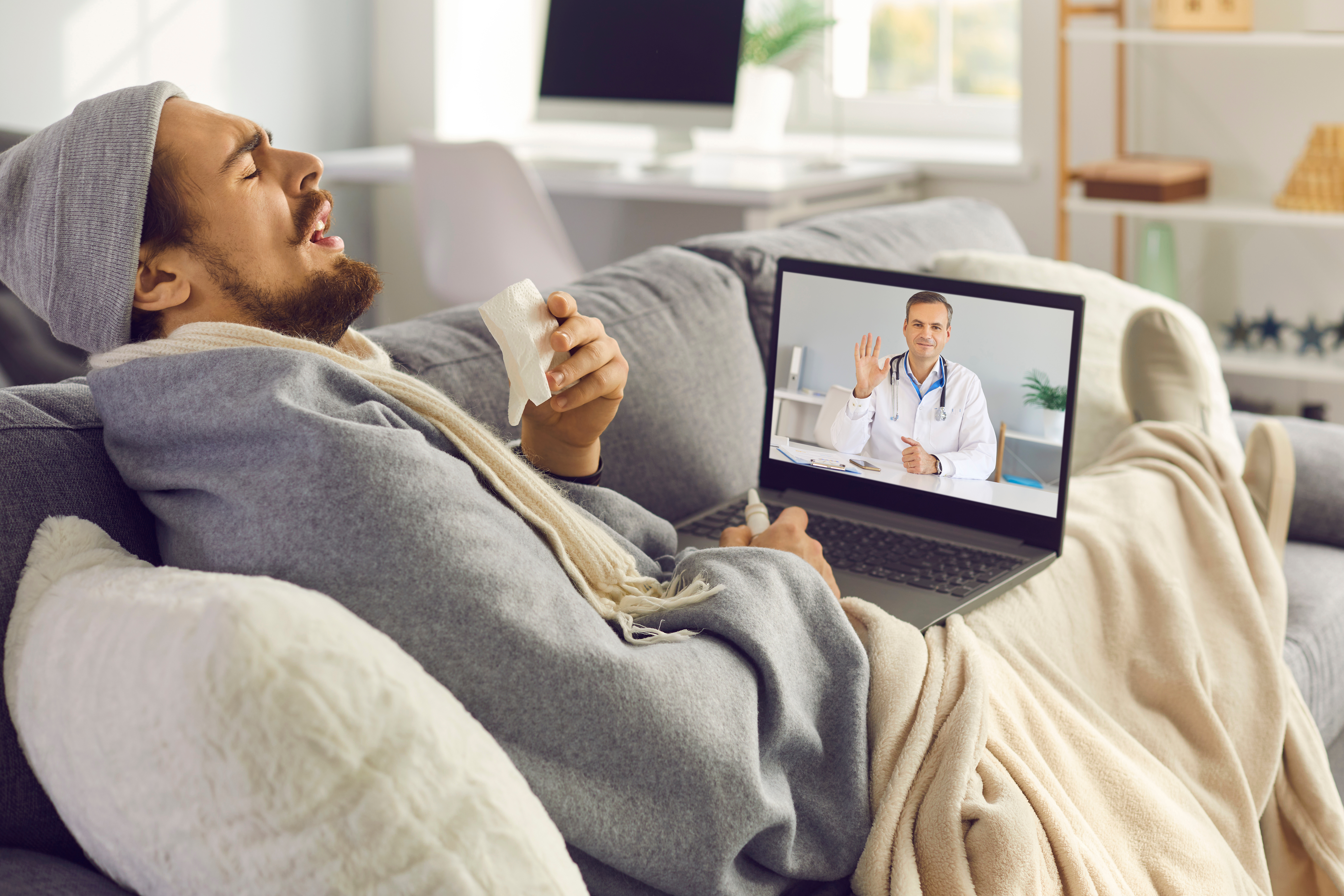 A man in a sweater, hat and blankets on his couch, mid-sneeze. He is holding a laptop where a doctor is waving at him from the screen.