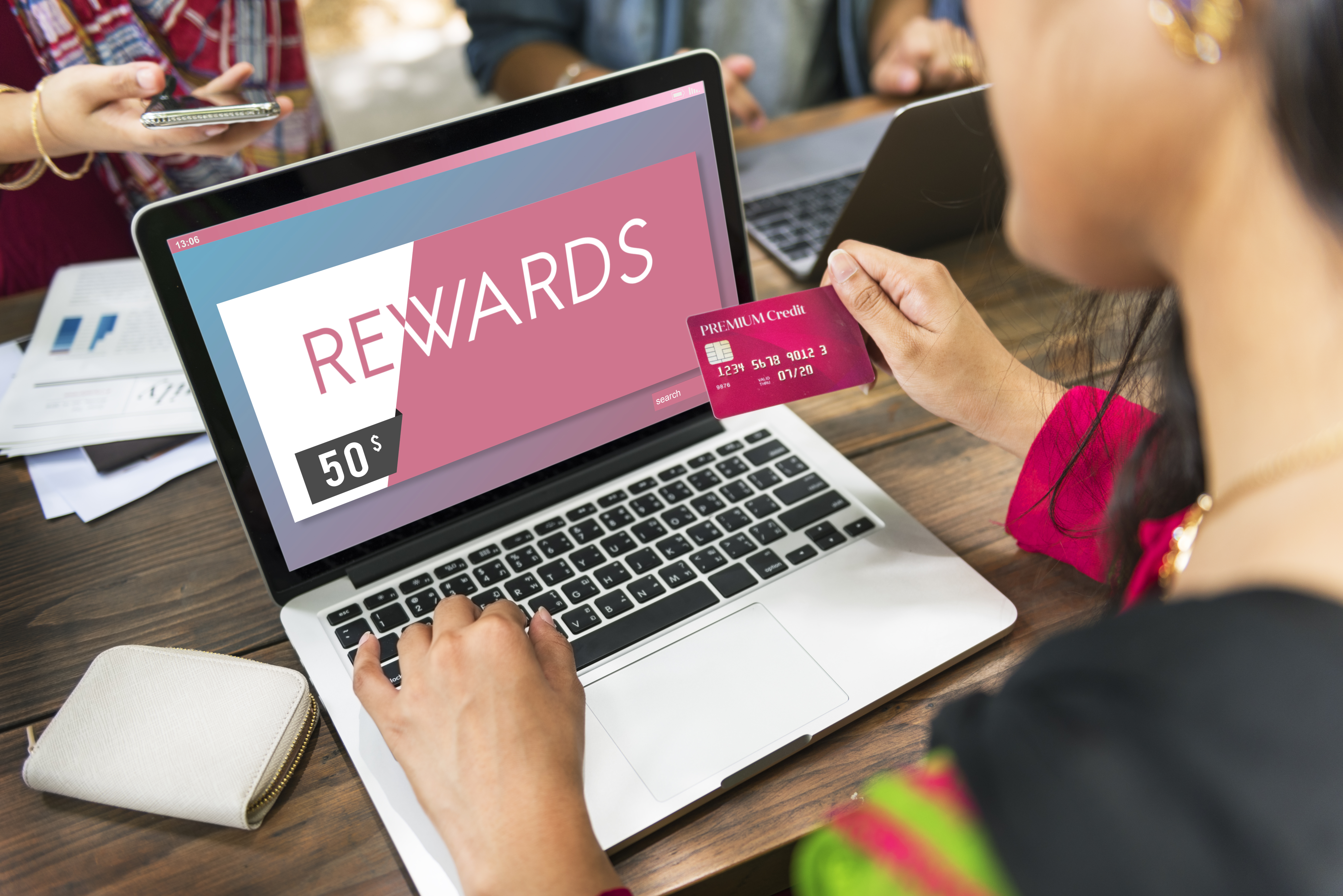 A woman sitting at a laptop, holding out her credit card to check the number. On the laptop is a page that says "REWARDS."