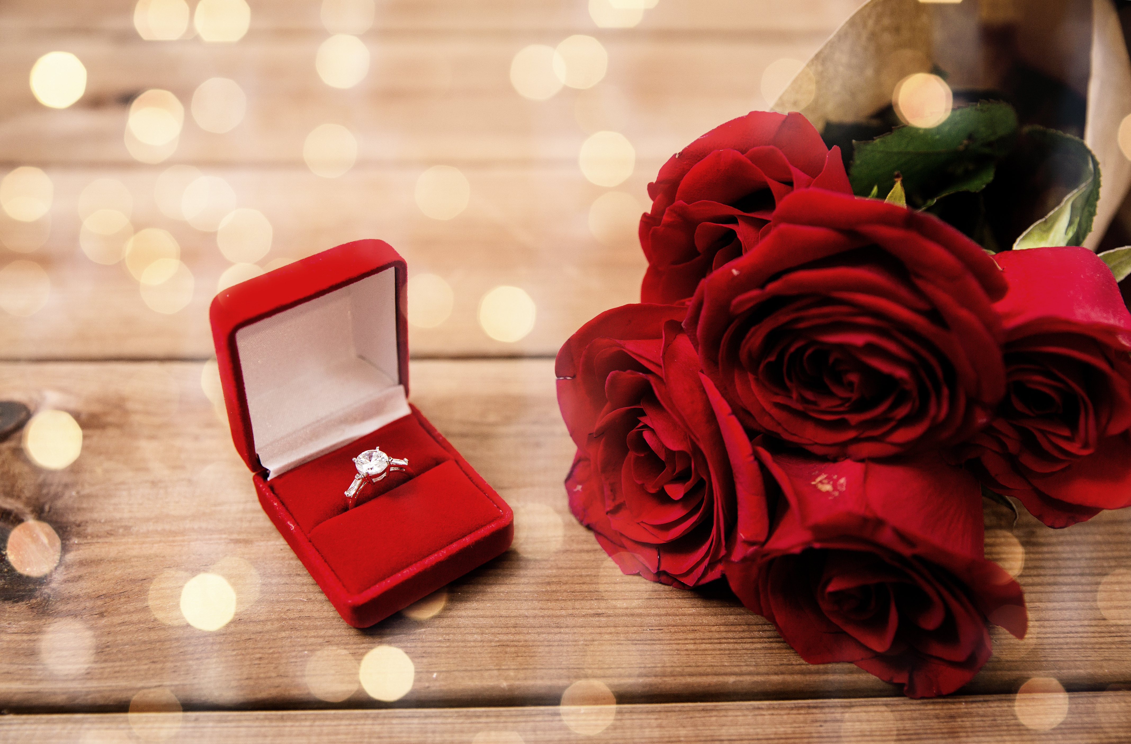 An engagement ring in a red box next to a bouquet of red roses on a wooden table.